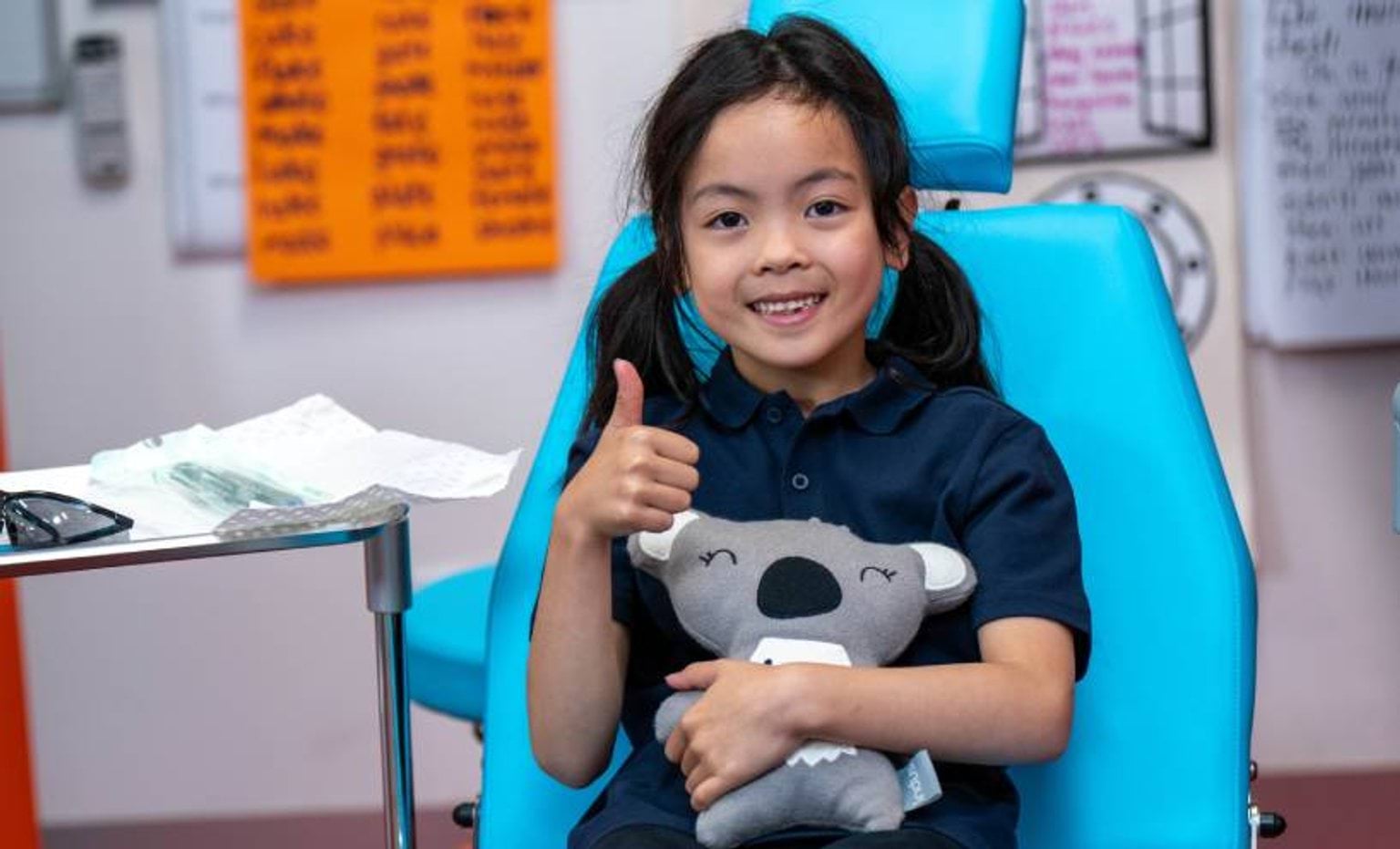 Girl in Smile Squad dental chair Girl sitting in a turquoise dentist chair giving a thumbs up and hugging a koala bear toy