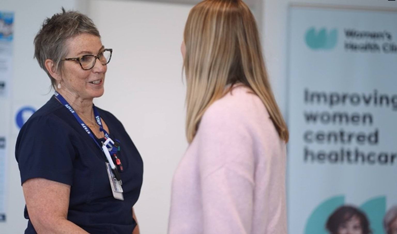 A nurse in navy scrubs speaking with a blond woman in a pink top.
